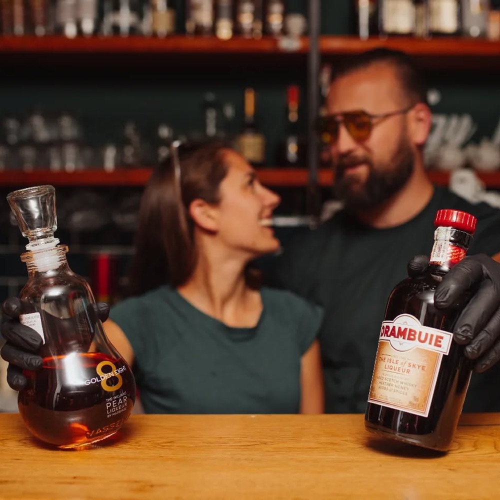 Két bartender mosolyogva mutatja az italos üvegeket a pult felett